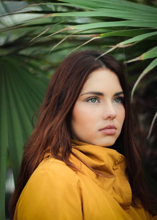 A young woman facing to her side with palm tree leaf in the background.